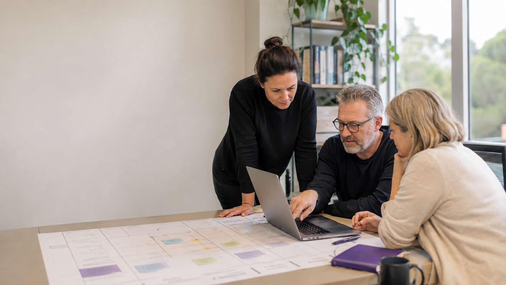 Three colleagues in a bright office reviewing a printed service blueprint and a laptop together